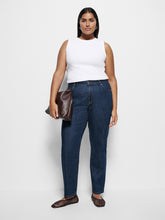 Load image into Gallery viewer, A woman modeling dark blue straight-leg jeans with a white sleeveless top, holding a brown leather clutch, and wearing brown woven flat shoes against a light gray studio background.
