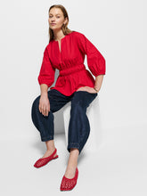Load image into Gallery viewer, Woman wearing a red blouse and blue jeans sitting on a white chair.
